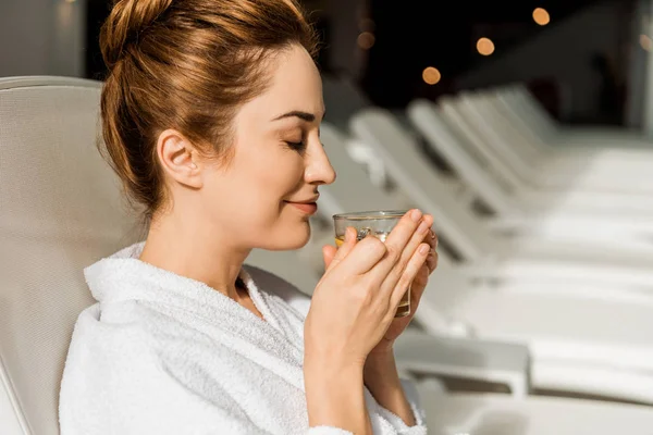 Side view of happy young woman holding cup of herbal drink while resting on sunbed in spa — Stock Photo