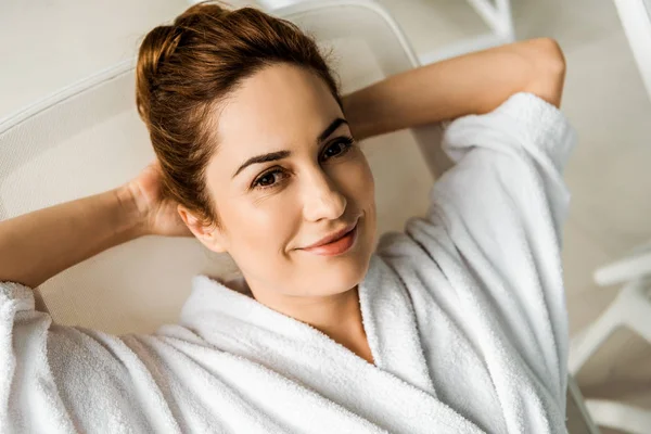 Hermosa mujer joven en albornoz acostado con las manos detrás de la cabeza y sonriendo a la cámara en el spa — Stock Photo