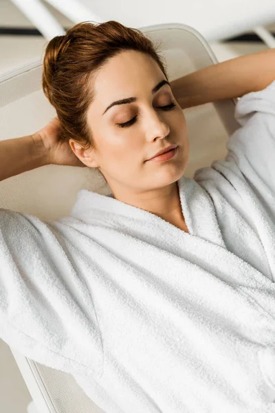 Atractiva joven en albornoz acostada con las manos detrás de la cabeza y los ojos cerrados en el spa — Stock Photo