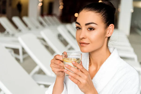 Привлекательная молодая женщина в халате пьет травяной чай и смотрит вдаль в спа — стоковое фото