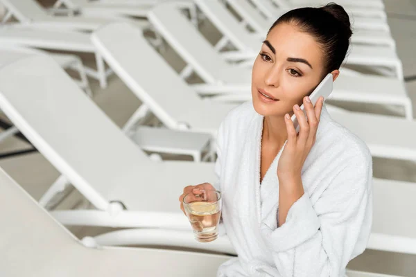 Hermosa mujer joven sosteniendo taza de té de hierbas y hablando por teléfono inteligente en el spa - foto de stock