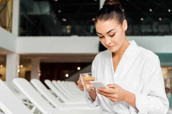 Young woman in bathrobe holding cup of herbal tea and using smartphone in spa — Stock Photo