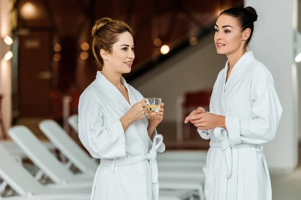 Smiling young women in bathrobes enjoying herbal tea and talking in spa — Stock Photo