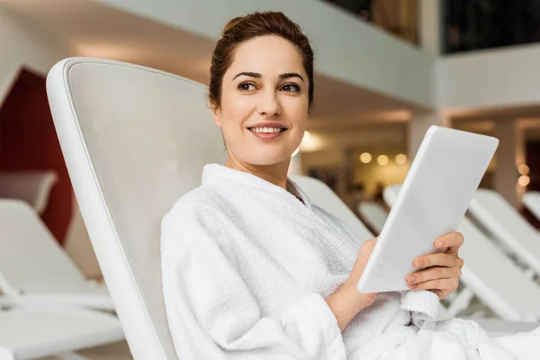 Mujer joven sonriente en albornoz usando tableta digital y mirando hacia otro lado mientras se relaja en el spa - foto de stock