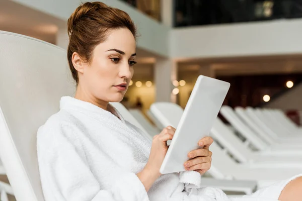 Приваблива молода жінка в халаті використовує цифровий планшет, розслабляючись у спа-центрі — стокове фото