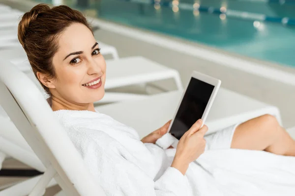 Молодая женщина в халате с помощью цифрового планшета и улыбаясь на камеру во время отдыха в спа — стоковое фото