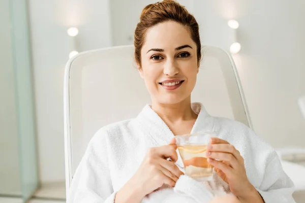 Junge Frau im Bademantel hält Tasse mit Kräutergetränk in der Hand und lächelt in die Kamera, während sie sich im Wellnessbereich entspannt — Stockfoto