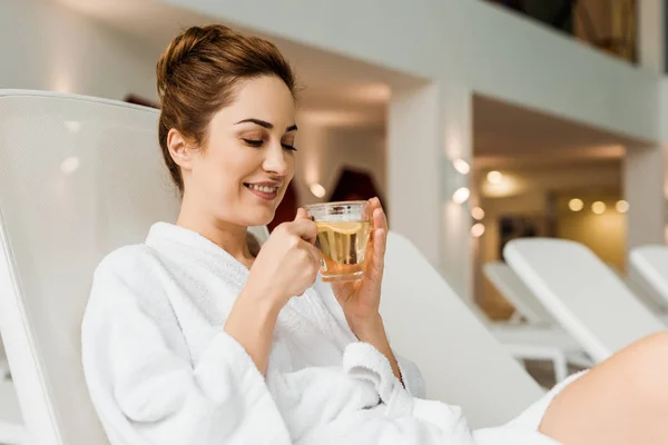 Sonriente joven mujer sosteniendo taza con bebida de hierbas mientras se relaja en el spa - foto de stock