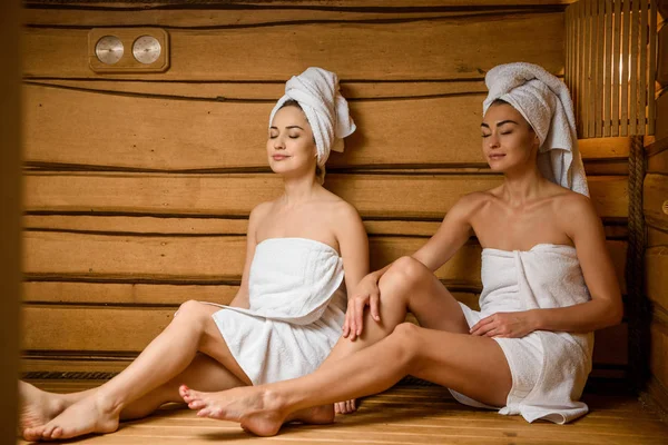 Hermosas mujeres jóvenes en toallas sentadas con los ojos cerrados en la sauna - foto de stock