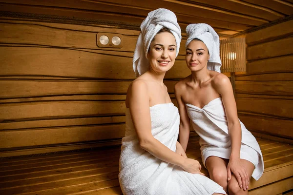 Attraenti giovani donne sedute insieme in sauna e sorridenti alla macchina fotografica — Foto stock