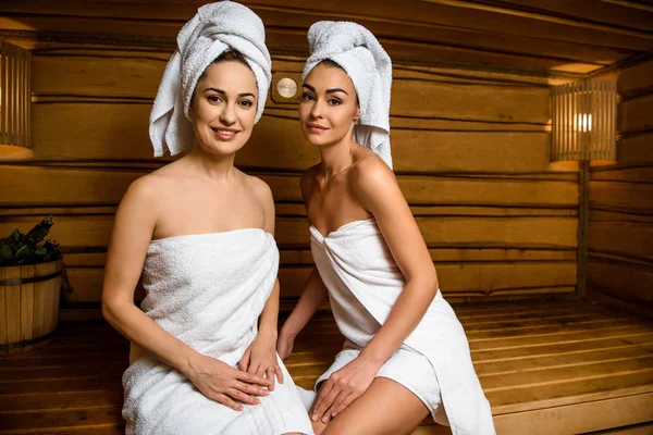 Hermosas mujeres jóvenes sentadas juntas en la sauna y sonriendo a la cámara - foto de stock