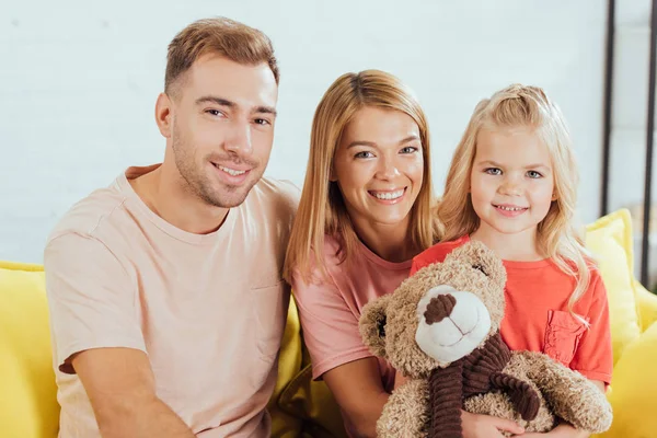 Lächelnde Eltern, die auf der Couch sitzen, in die Kamera schauen und ihre Tochter einen Teddybär in der Hand halten — Stockfoto