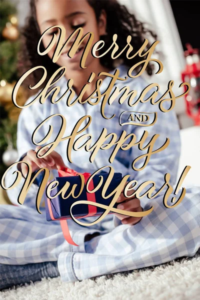 Focus selettivo di sorridente adorabile bambino afroamericano in pigiama che apre il regalo di Natale a casa con scritte 