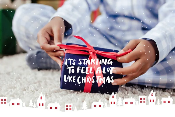 Imagen recortada de niño afroamericano en pijama abriendo regalo de Navidad en el suelo con letras, nieve e ilustración de casas 