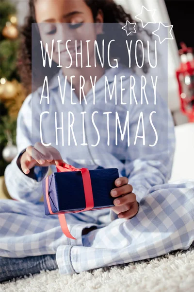 Вибірковий фокус усміхненої чарівної афроамериканської дитини в піжамі, що відкриває різдвяний подарунок вдома з написом 