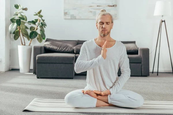 Homem adulto com olhos fechados meditando na posição de lótus no tapete de ioga em casa — Fotografia de Stock