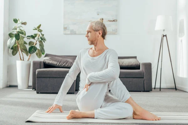 Concentrato uomo adulto seduto in posa yoga sul tappeto e guardando lontano a casa — Foto stock