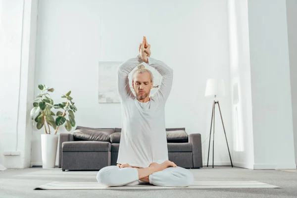Hombre adulto meditando en posición de loto con mudra signo en esterilla de yoga en casa - foto de stock
