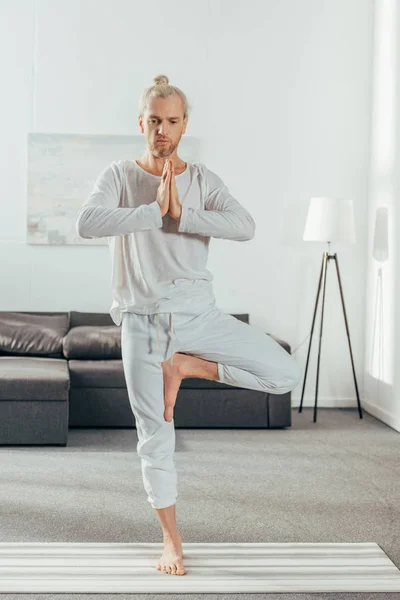 Uomo adulto in piedi in posa albero con gesto namaste sul tappetino yoga a casa — Foto stock