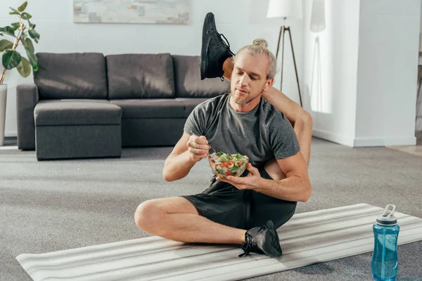 Hombre atlético flexible con la pierna detrás de la cabeza sentado en la estera de yoga y comer ensalada de verduras - foto de stock