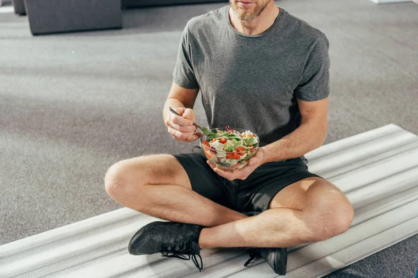 Tiro recortado de hombre atlético en ropa deportiva sentado en la esterilla de yoga y tazón de celebración con ensalada de verduras en casa - foto de stock