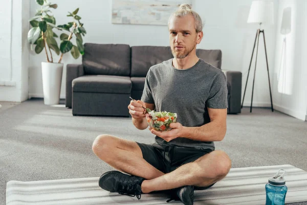 Sportlicher erwachsener Mann in Sportbekleidung mit Schüssel Gemüsesalat und Blick in die Kamera, während er zu Hause auf der Yogamatte sitzt — Stockfoto