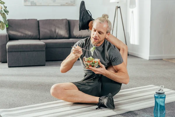 Гнучкий дорослий чоловік з ногою позаду голови сидить на килимку для йоги та їсть овочевий салат — стокове фото