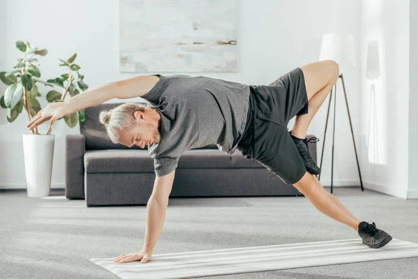 Homme athlétique flexible en vêtements de sport pratiquant planche latérale sur tapis de yoga à la maison — Photo de stock