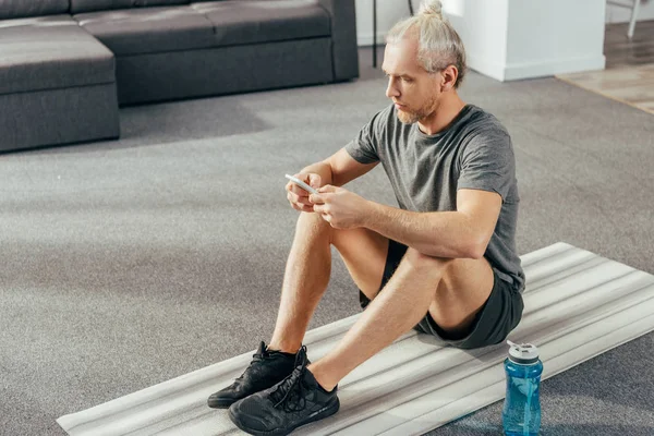 Hochwinkelaufnahme eines erwachsenen Mannes in Sportbekleidung, der in einer Yogamatte sitzt und zu Hause das Smartphone benutzt — Stockfoto