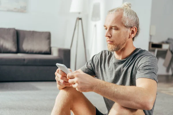 Ernstzunehmender erwachsener Mann in Sportbekleidung sitzt und Smartphone zu Hause benutzt — Stockfoto