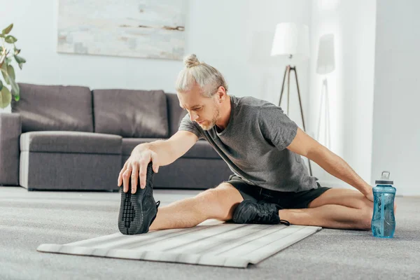 Athletischer Mann in Sportbekleidung sitzt auf Yogamatte und dehnt sich zu Hause — Stockfoto