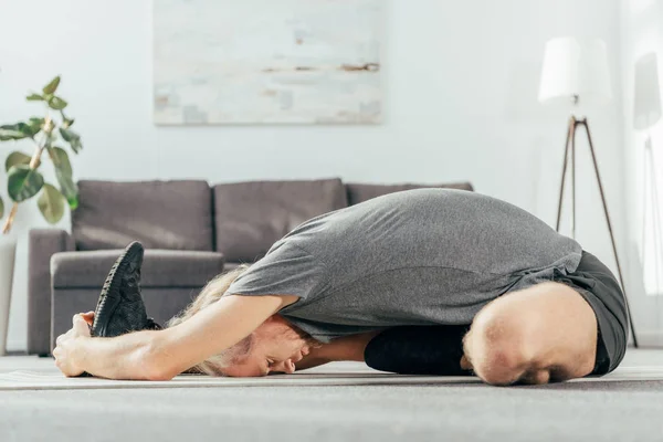Vista lateral del hombre atlético en ropa deportiva haciendo ejercicio y estirándose en la esterilla de yoga en casa - foto de stock