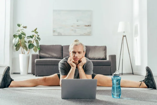 Desportivo adulto homem fazendo split no tapete de ioga e olhando para laptop — Fotografia de Stock
