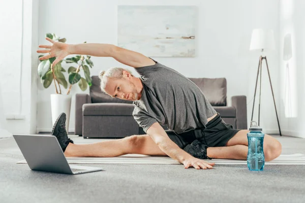 Спортивний дорослий чоловік тягнеться на йога килимок і дивиться на ноутбук — стокове фото