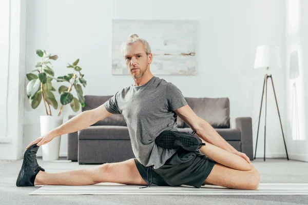 Hombre adulto guapo estirándose en la estera de yoga y mirando a la cámara mientras hace ejercicio en casa - foto de stock