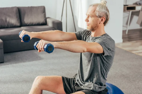 Sportlicher erwachsener Mann sitzt auf fittem Ball und trainiert zu Hause mit Kurzhanteln — Stockfoto