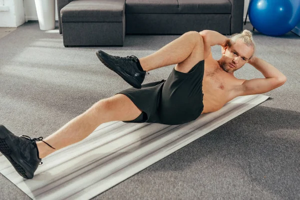 Hombre musculoso sin camisa haciendo ejercicios abdominales en la esterilla de yoga en casa - foto de stock