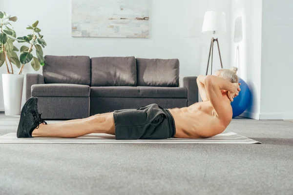 Vista lateral del hombre sin camisa haciendo ejercicios abdominales en la esterilla de yoga en casa - foto de stock