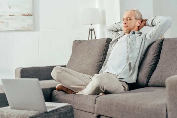 Handsome behind man sitting with hands behind head on couch and looking at laptop — Stock Photo