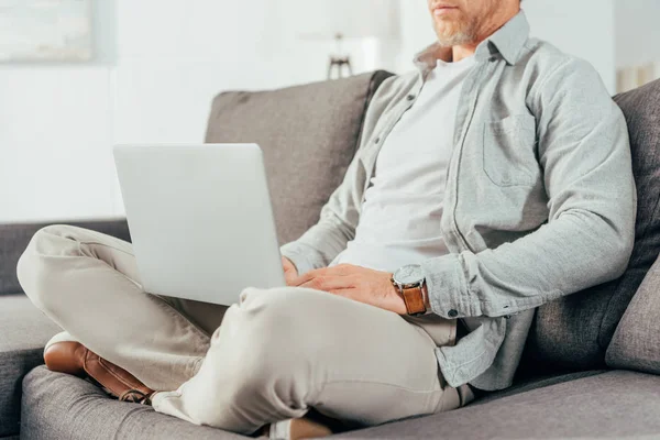 Tiro cortado de homem sentado no sofá e usando laptop — Fotografia de Stock