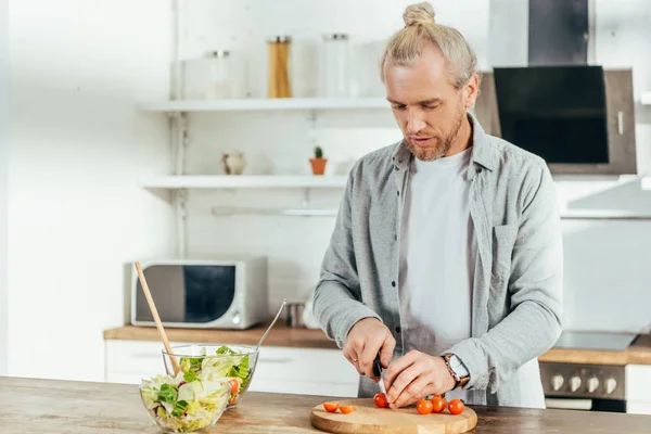 Homem adulto bonito cortando tomates cereja e cozinhar salada de legumes na cozinha — Fotografia de Stock