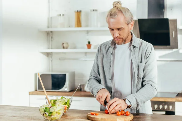 Handsome adult man chopping cherry tomatoes and cooking vegetable salad in kitchen — Stock Photo