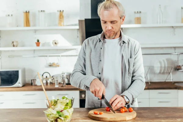 Homme hachant tomates cerises et cuisine salade de légumes dans la cuisine — Photo de stock
