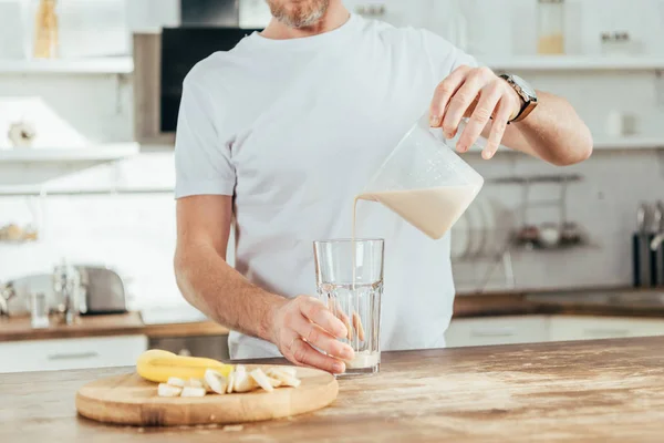 Tiro cortado de homem adulto derramando batido de proteína de banana em vidro em casa — Fotografia de Stock