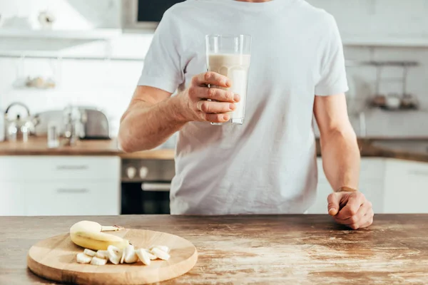 Tiro cortado de homem atlético segurando vidro com batido de proteína de banana em casa — Fotografia de Stock