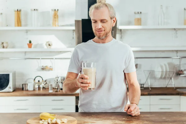 Uomo sportivo adulto sorridente che tiene il vetro con frullato di proteine della banana a casa — Foto stock