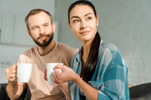 Beautiful husband and wife sitting on couch drinking coffee and looking away — Stock Photo