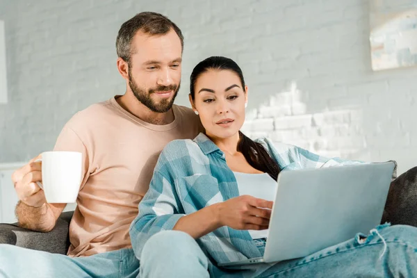 Marido sosteniendo la taza de café mientras que la esposa hermosa usando el ordenador portátil en el sofá - foto de stock