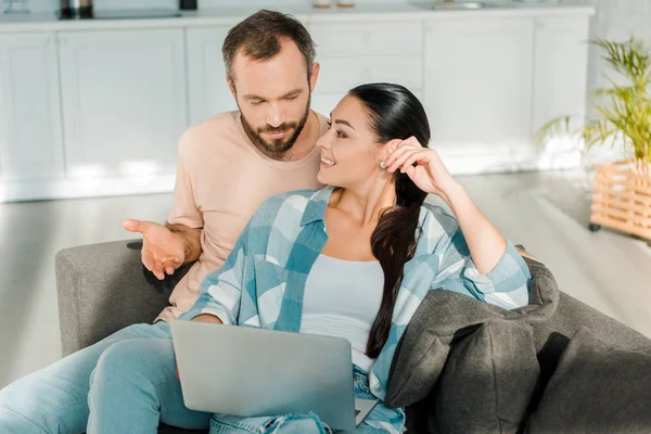 Marido guapo y esposa sonriente sentado en el sofá y el uso de la computadora portátil en casa - foto de stock