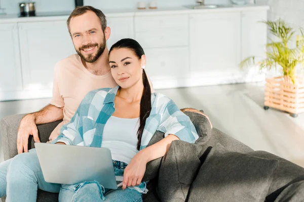 Husband and attractive wife sitting on couch, looking at camera and using laptop at home — Stock Photo